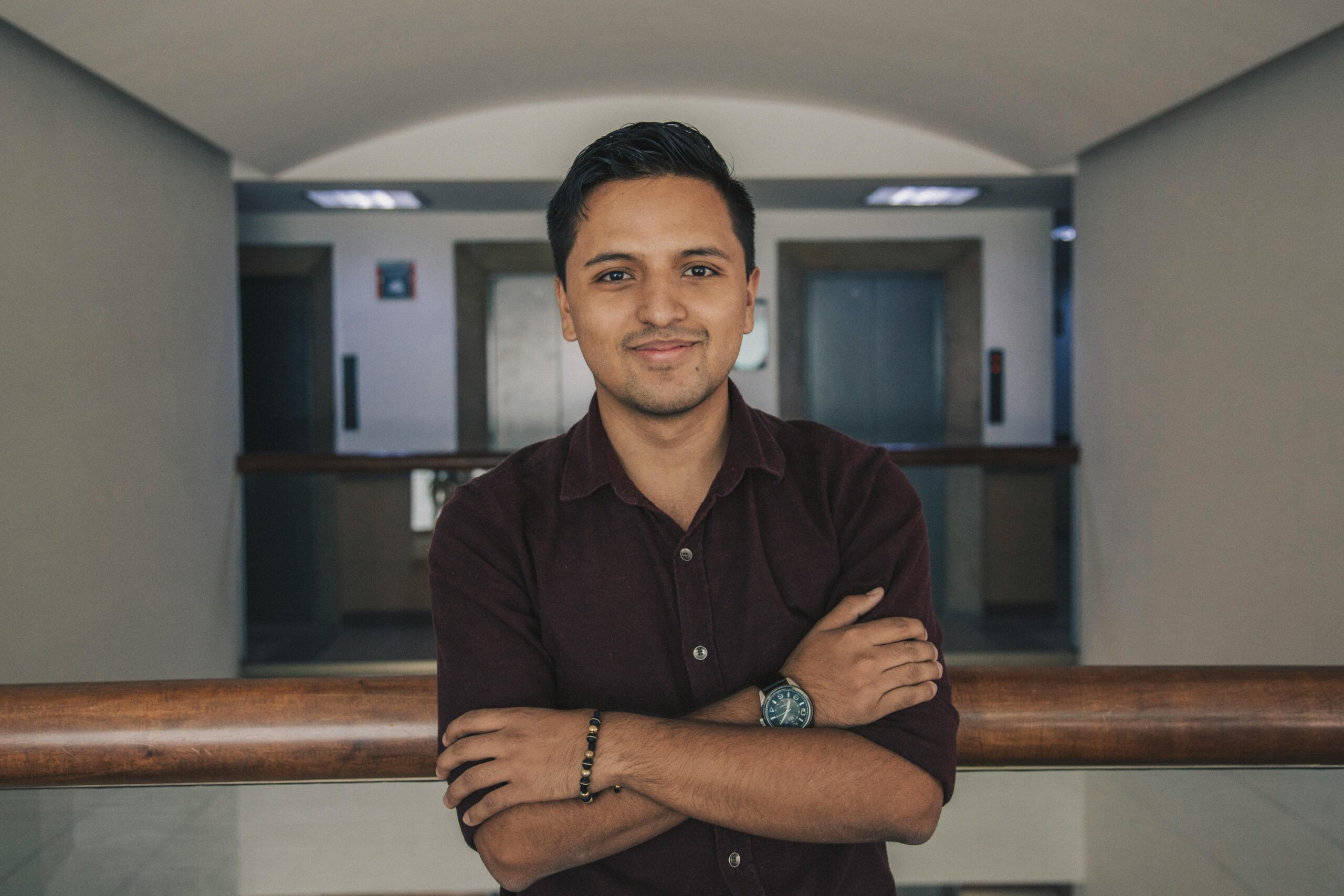 Positive Indian in casual outfit standing with crossed arms near glass fence while smiling and looking at camera in modern corridor