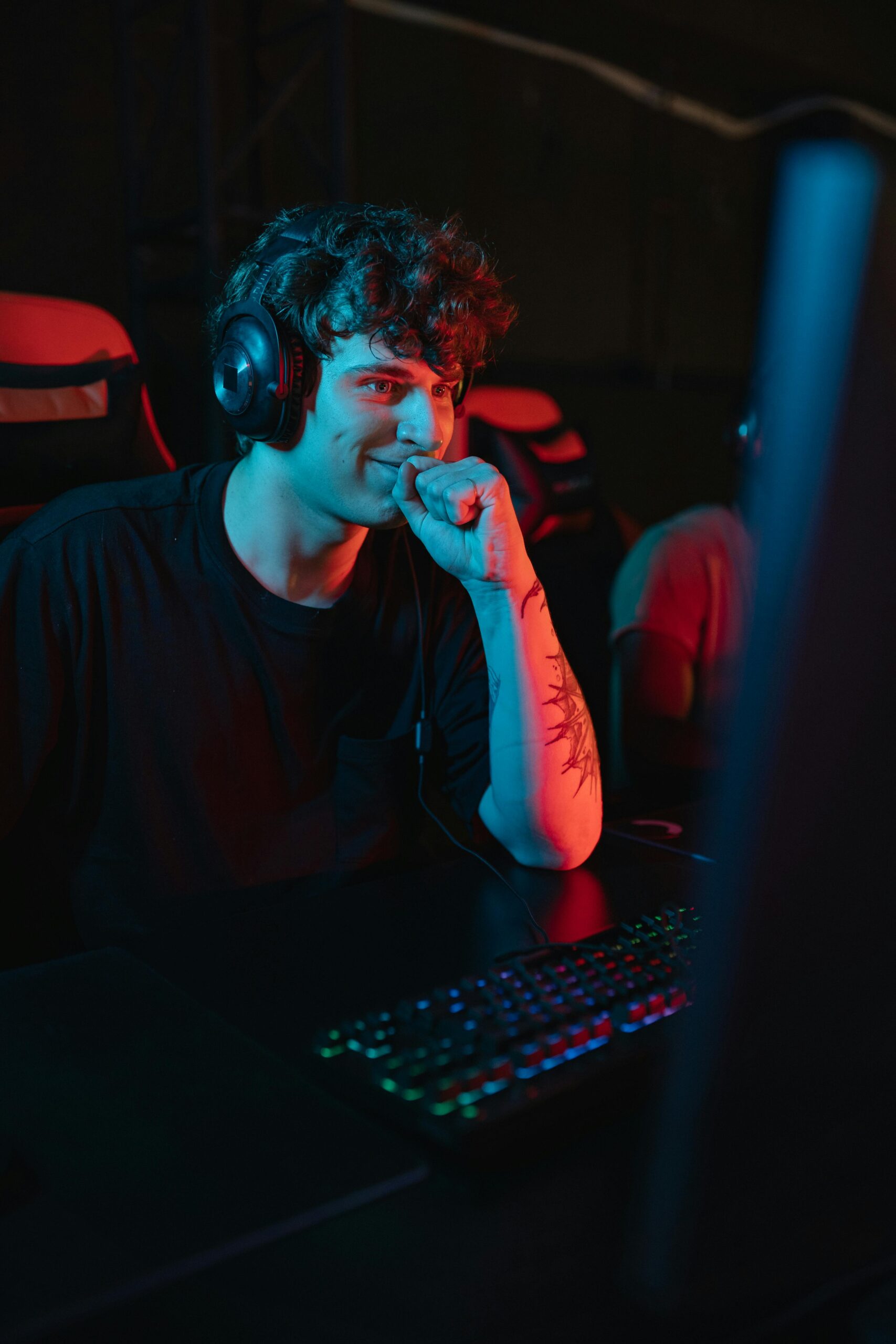 A young man sitting and gaming indoors, wearing a headset with a focused expression under colorful lighting.